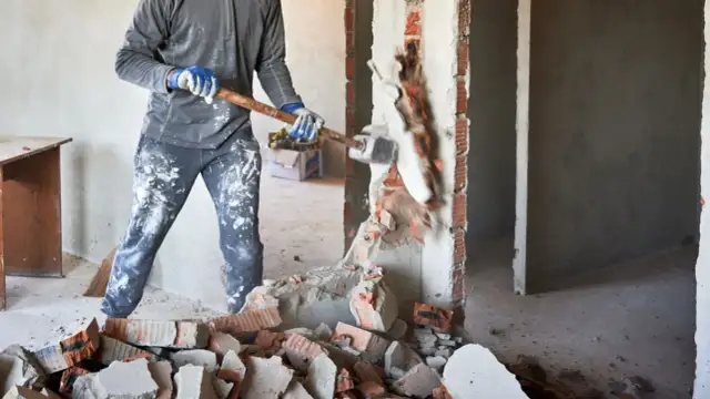 Mężczyzna w rękawicach i ubraniu w plamach farby burzy ścianę młotem. Czy spółdzielnia może zabronić remontu mieszkania?