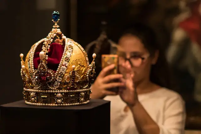 Złota korona, ozdobiona perłami i rubinami, przypomina wielki mogoł diament. Kobieta w okularach robi jej zdjęcie telefonem.