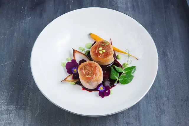Eleganckie danie z dwóch medalionów mięsa zawiniętych w boczek, podane z purée, marchewką i kwiatami.