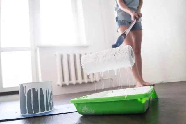 Osoba maluje wałkiem białą farbą, która kapie do zielonej kuwety.