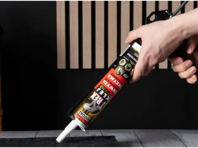 Dłoń trzymająca pistolet z tubą kleju montażowego na tle paneli lamelowych.