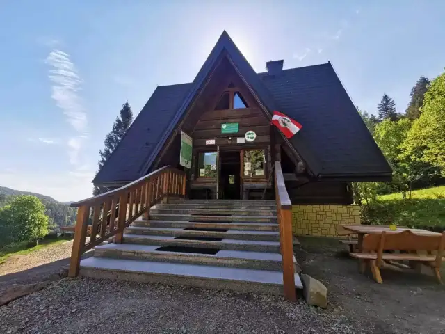 Drewniana bacówka pod honem, z kamiennymi schodami i drewnianymi ławami, otoczona zielenią.