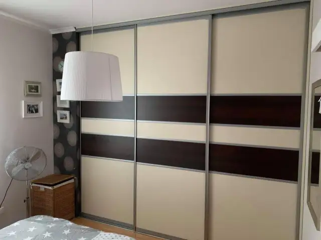 Oklejanie szafy przesuwnej w beżowo-brązowe pasy. W tle widać lampę, zdjęcia i wentylator.