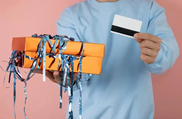 Osoba w niebieskiej bluzie trzyma pomarańczowe pudełko prezentowe z niebieskimi wstążkami i białą kartę.