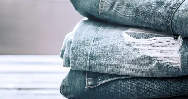 Stos znoszonych dżinsów w odcieniu **kolor denim**. Widać przetarcia i postrzępione fragmenty materiału, świadczące o historii noszenia.