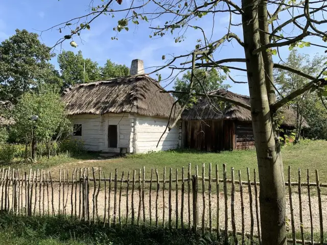 Drewniane chaty kryte strzechą w skansenie, przed nimi płotek z gałęzi.