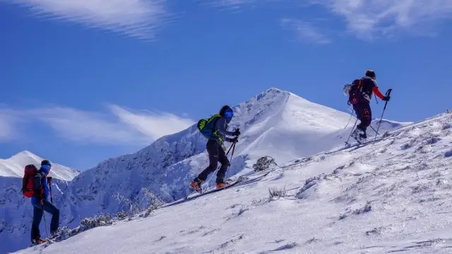 Trzy osoby na nartach skiturowych wspinają się po ośnieżonym stoku. W tle majestatyczne, pokryte śniegiem góry i błękitne niebo.