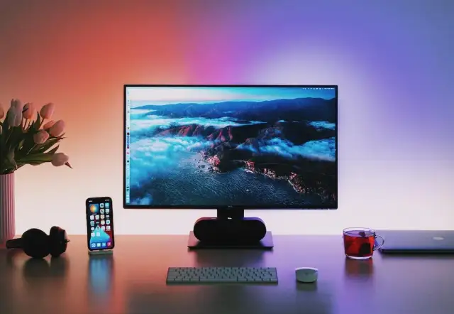 Biurko z monitorem wyświetlającym krajobraz, telefonem, słuchawkami i kubkiem herbaty.