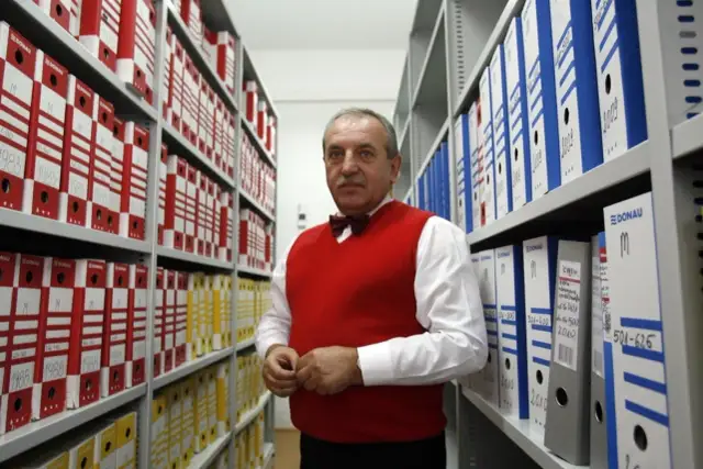 Starszy mężczyzna w czerwonym swetrze stoi między regałami pełnymi segregatorów.