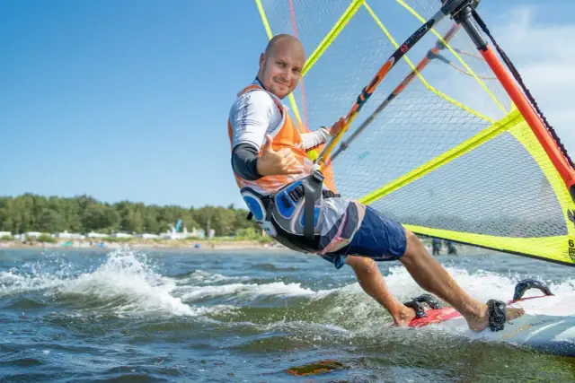Mężczyzna na desce windsurfingowej, uśmiecha się i pokazuje kciuk w górę.