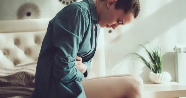 Kobieta w niebieskiej koszuli trzyma się za brzuch, cierpiąc z powodu bólu. Zastanawia się, ile trwa okres i jak sobie z nim poradzić.