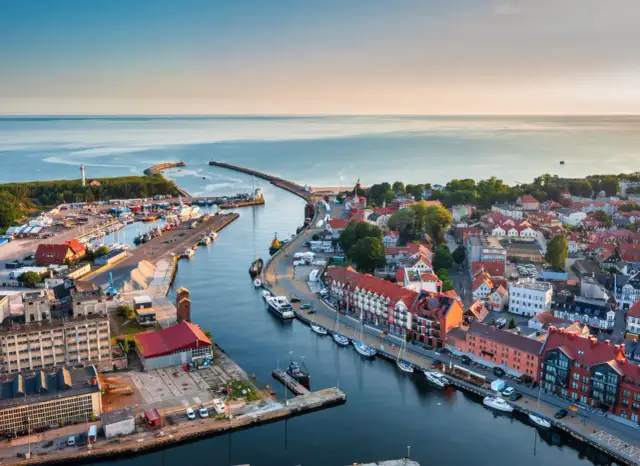 Widok z lotu ptaka na port w Ustce z łodziami, budynkami i morzem w tle.