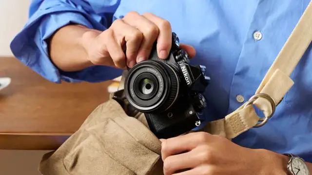 Osoba w niebieskiej koszuli wkłada czarny aparat Nikon z obiektywem do torby.
