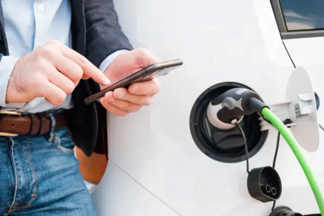 Mężczyzna sprawdza na telefonie, jak rozliczyć ładowanie samochodu elektrycznego w domu. Zielony kabel podłączony do pojazdu.