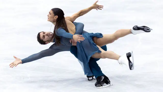 Para tańczy na lodzie w błękitnych strojach, wykonując skomplikowaną figurę. Ich ruchy to czysta jazda figurowa na lodzie.