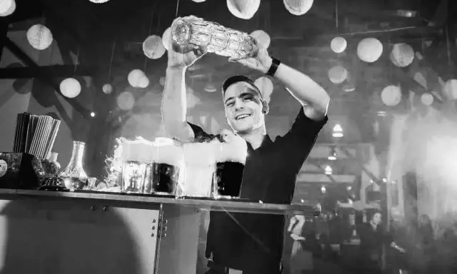 Uśmiechnięty barman z shakerem nad głową, przygotowuje drinki z dymem.