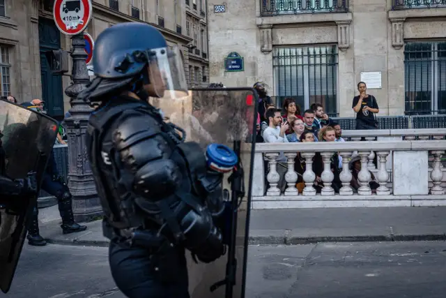 Policjant w pełnym rynsztunku ochronnym z tarczą, obserwuje tłum za barierką.
