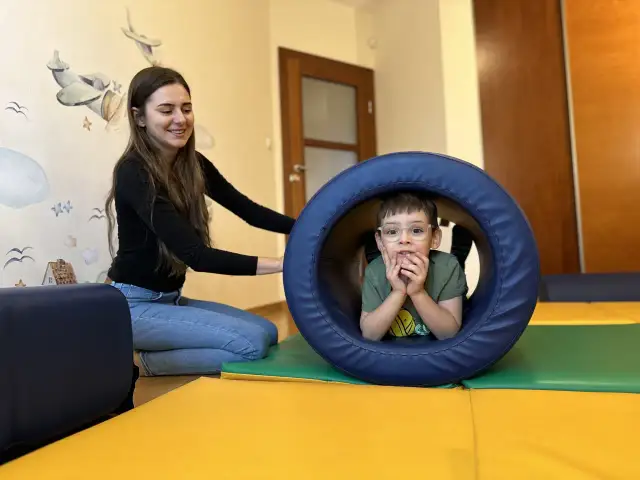 Chłopiec w okularach w tunelu terapeutycznym, obok uśmiechnięta terapeutka.