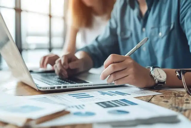 Osoba w niebieskiej koszuli pisze piórem na dokumentach z wykresami obok laptopa.