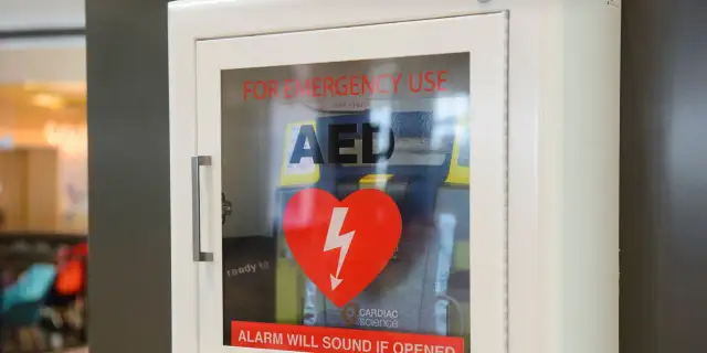 Biała szafka z defibrylatorem AED, czerwonym sercem z błyskawicą i napisem "FOR EMERGENCY USE".