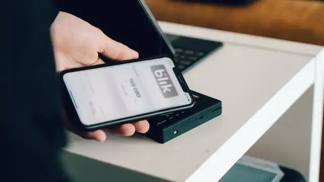 Dłoń trzyma smartfon z aplikacją Blik i kodem transakcji nad terminalem płatniczym.