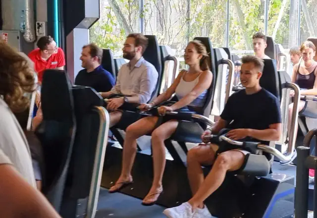 Ludzie siedzą w wagonikach kolejki górskiej, niektórzy uśmiechnięci, inni patrzą przed siebie.