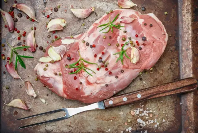 Surowe mięso jagnięce, czosnek, rozmaryn i kolorowy pieprz na blaszce, gotowe do peklowania na mokro. Widelec obok.