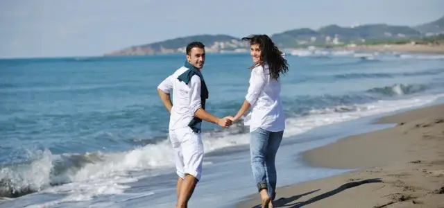 Para na plaży, trzymając się za ręce, cieszy się majówką. Słońce, morze i góry w tle tworzą idealny krajobraz.