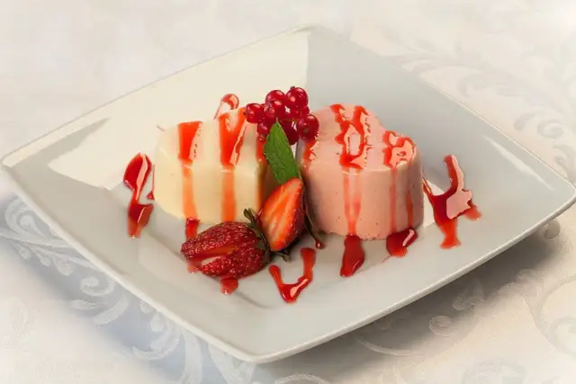 Dwie panna cotta w kształcie serc, polane sosem truskawkowym, ozdobione owocami i miętą.