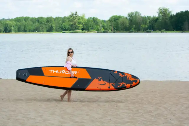 Kobieta z pomarańczowo-czarnymi deskami do pływania z wiosłem na piaszczystej plaży, gotowa do wodnych przygód.