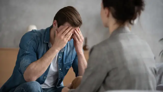 Mężczyzna w dżinsowej koszuli z rękami na twarzy, siedzący naprzeciwko drugiej osoby.