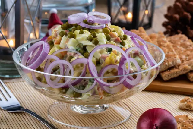 Pyszna sałatka jarzynowa, idealna na Wielkanoc, ozdobiona krążkami czerwonej cebuli. W tle widać świece i pieczywo.