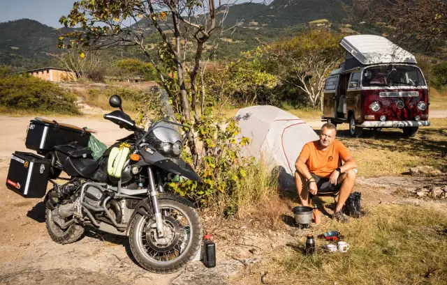 Motocykl, namiot, kamper i mężczyzna siedzący przy ognisku na tle gór.