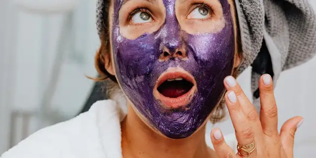 Kobieta z fioletową, błyszczącą maską na twarzy, w białym szlafroku i z ręcznikiem na głowie, cieszy się domowymi zabiegami pielęgnacyjnymi na twarz.