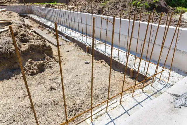Budowa fundamentów: pionowe pręty zbrojeniowe czekają na beton. Widać też białe bloczki fundamentowe i piasek.