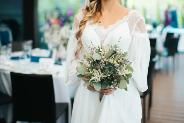 Panna młoda trzyma bukiet ślubny z czerwca, pełen zieleni, ostów i lawendy.