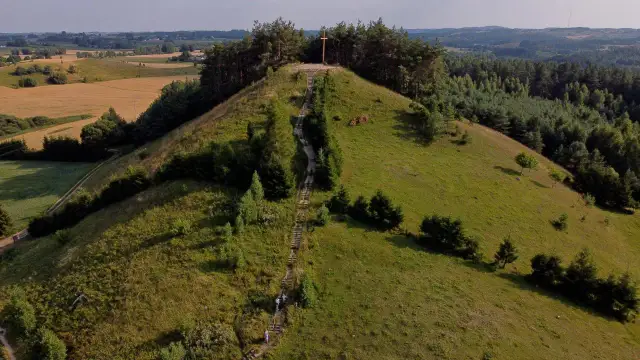 Wzgórze z drewnianym krzyżem na szczycie, schody prowadzące na górę. W oddali pola i lasy, krajobraz przypominający rezerwat przyrody cisowa.