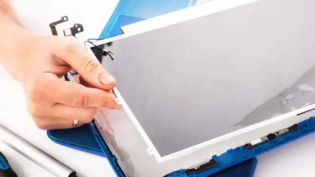 Dłoń trzyma matrycę w laptopie podczas naprawy. Widać narzędzia i niebieską obudowę.