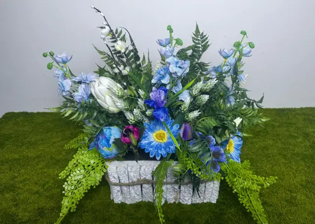 Sztuczna kompozycja kwiatowa w białym, kamiennym wazonie, na zielonym tle.