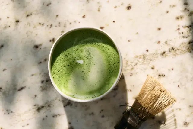 Zielone matcha w filiżance obok bambusowej miotełki na marmurowym blacie.