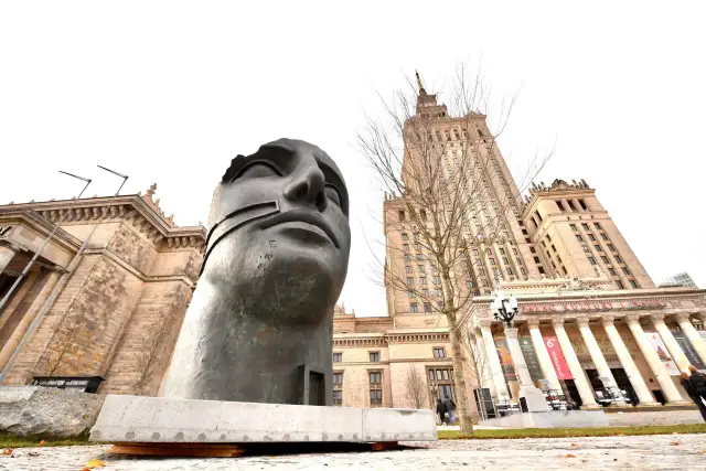 Monumentalna rzeźba głowy na tle Pałacu Kultury w Warszawie.