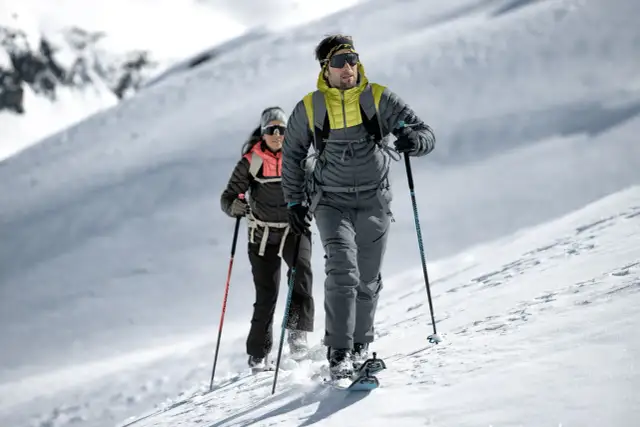 Dwóch narciarzy w kurtkach zimowych wspina się po ośnieżonym stoku. Jakie kurtki narciarskie wybrać na taką wyprawę?