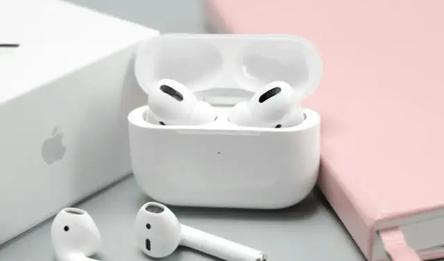 Białe słuchawki AirPods w etui, obok pudełka Apple i różowego notatnika.
