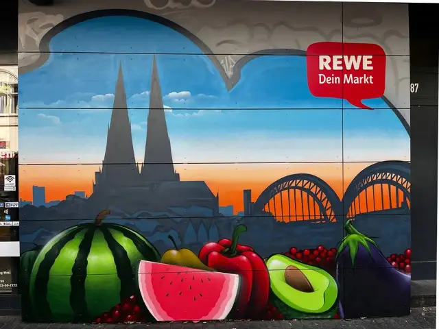 Graffiti mit Kölner Dom, Brücke und Früchten vor REWE-Logo.