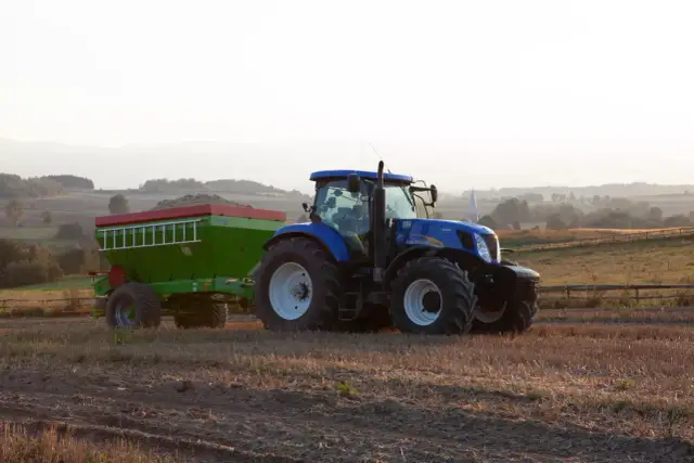 Niebieski traktor z przyczepą nawozową, gotowy do pracy na polu. Rolnik zastanawia się, jakie wapno na dofinansowanie będzie najlepsze.