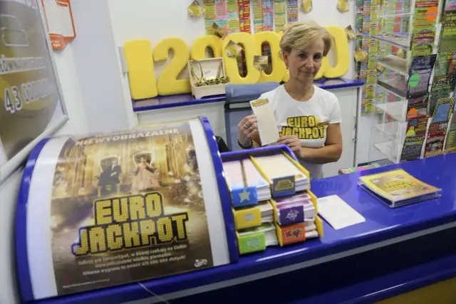 Kobieta w koszulce Eurojackpot trzyma kupon na tle stoiska z grami losowymi.