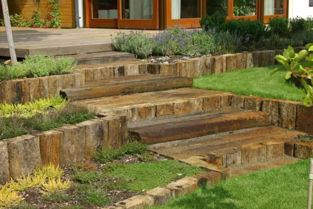 Drewniane schody prowadzące na taras na skarpie, otoczone zielenią i kwiatami.