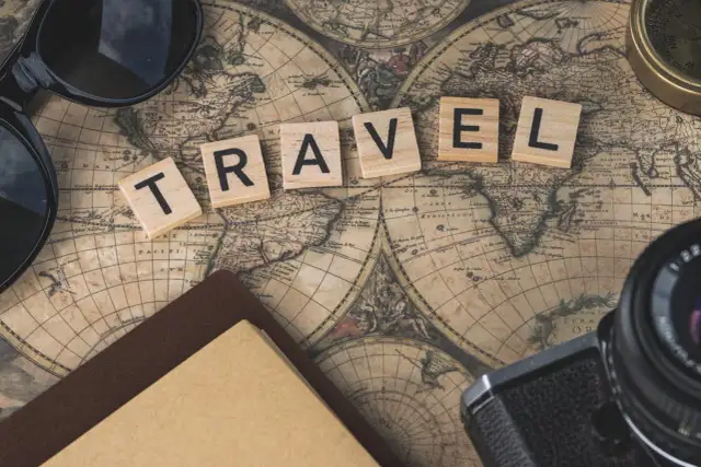 Drewniane klocki z napisem TRAVEL na starej mapie świata, obok okulary, aparat i kompas.
