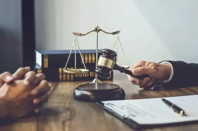 Dłoń trzymająca młotek sędziowski, waga sprawiedliwości i akta na drewnianym stole.