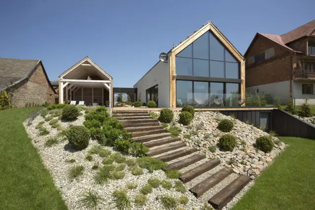 Nowoczesny dom z tarasem i drewnianymi schodami prowadzącymi na skarpę w ogrodzie. Inspiracje dla miłośników zieleni i architektury.